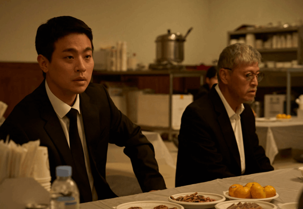 상복을 입은 남자들이 식탁 앞에 앉아 엄숙한 분위기를 보이는 장면