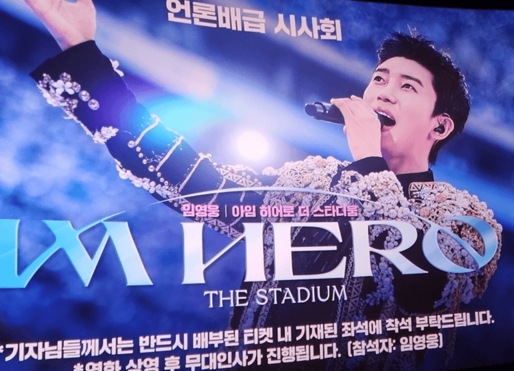 "화려한 장식이 있는 무대 의상을 입고 마이크를 들고 노래하는 임영웅, 뒤로 ‘IM HERO THE STADIUM’ 문구가 보이는 콘서트 홍보 이미지"